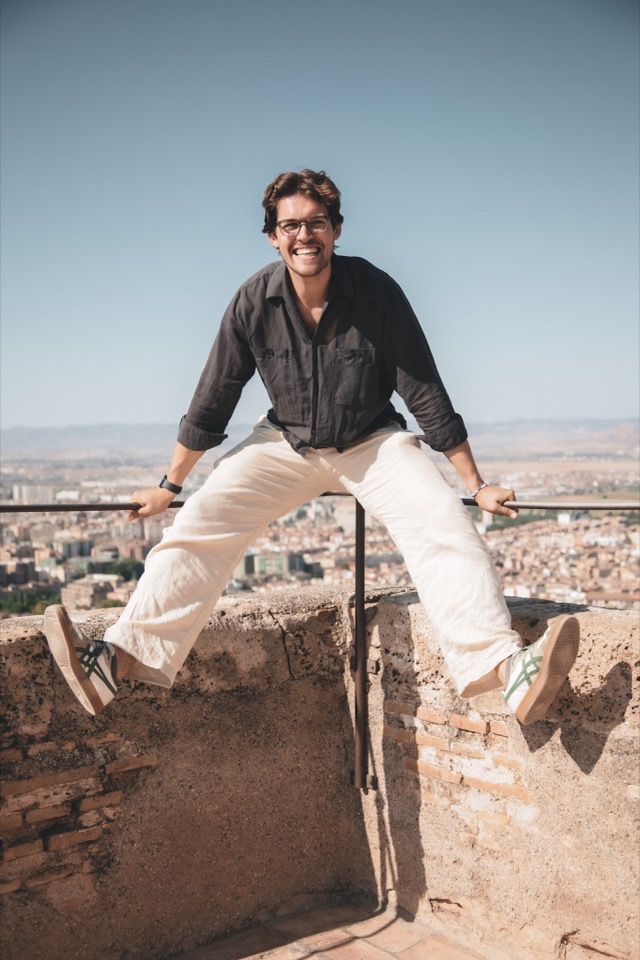 Felix sitting on a stone parapet above a sunlit city, arms resting on a metal rail, hills behind him.