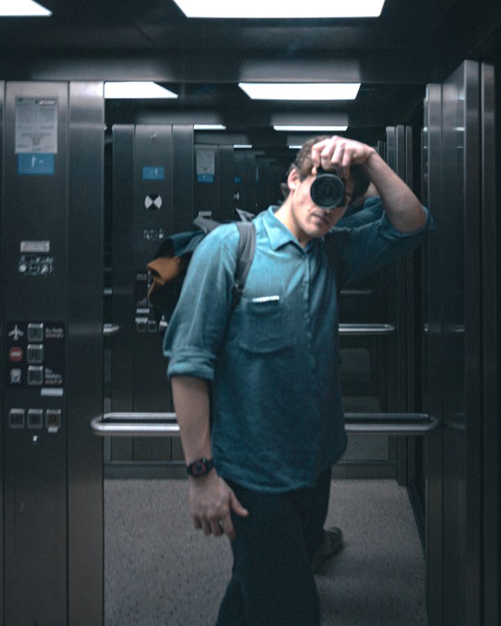 Felix in a lift, holding a camera up to his eye, reflected in the mirrored walls.