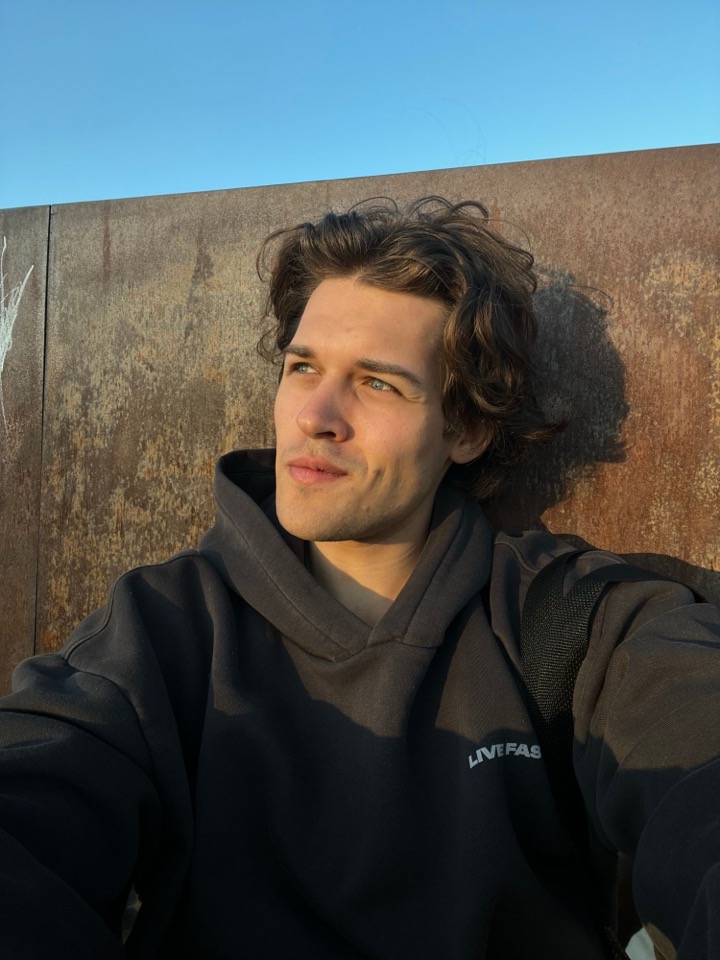 Felix outdoors in a dark hooded sweatshirt, looking away from the camera, with a weathered steel wall and a clear sky behind him.