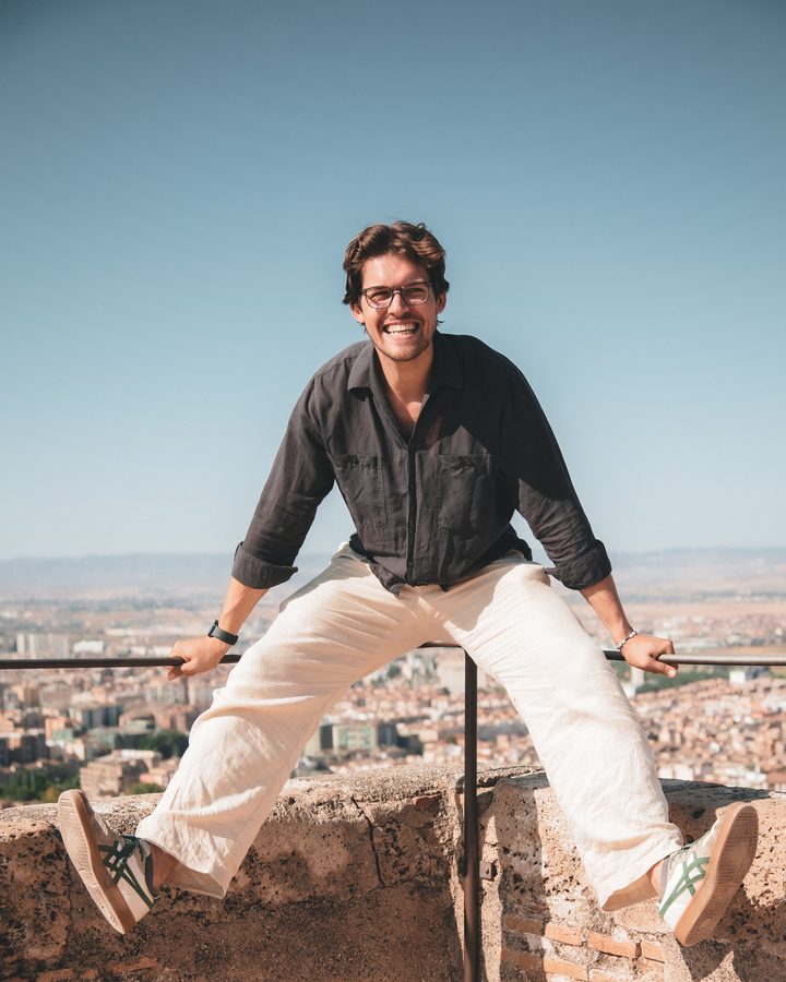 Felix balancing on a rampart above a sunlit city, laughing, glasses on.
