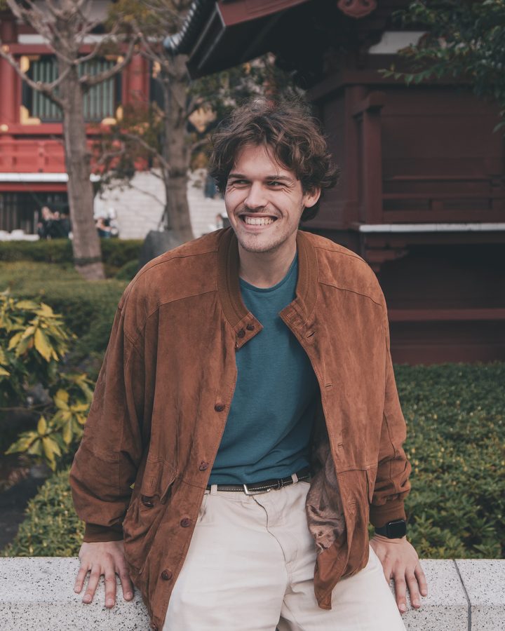 Felix standing in front of a temple in Tokyo, tan suede jacket, trees and a tiled roof behind him.